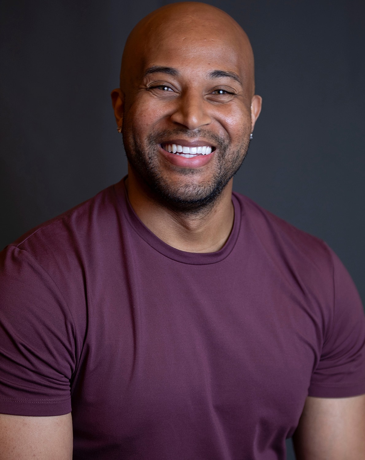 Smiling man wearing a maroon shirt, dark background.